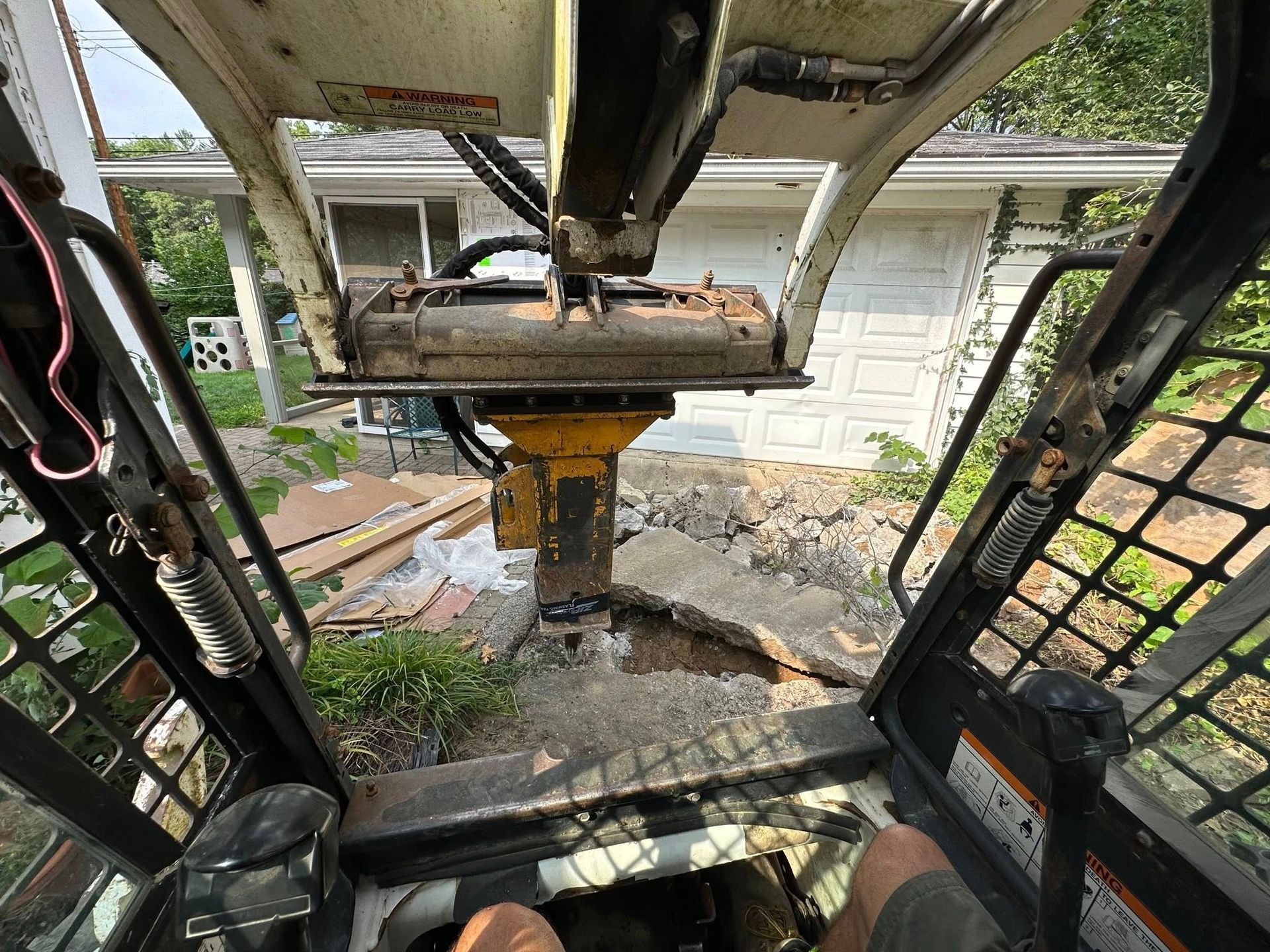Interior view from a damaged excavator cab with rusted controls, looking out toward a house and yard.