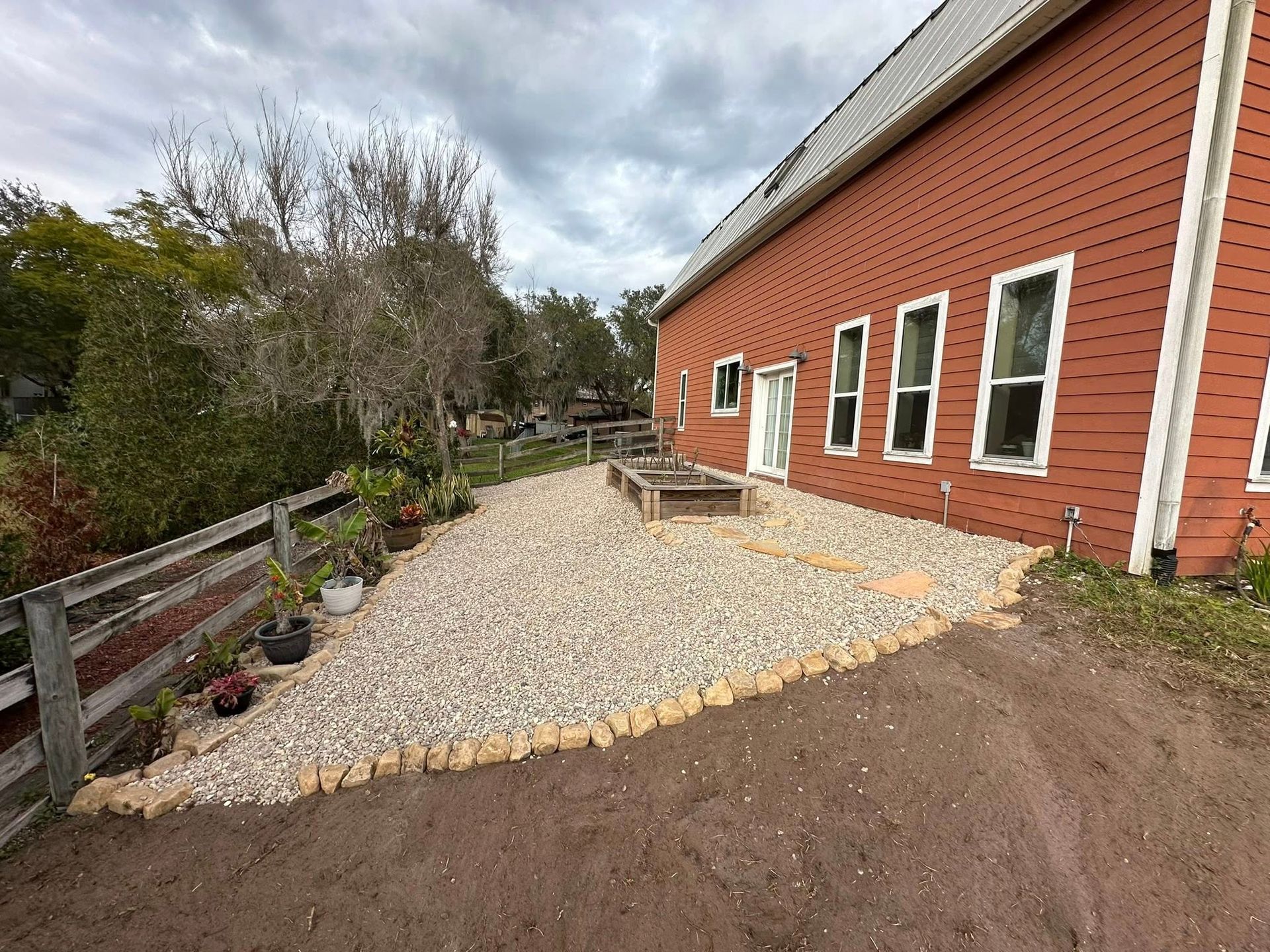 Gravel patio beside a red brick house with windows, wooden deck, and cloudy sky.
