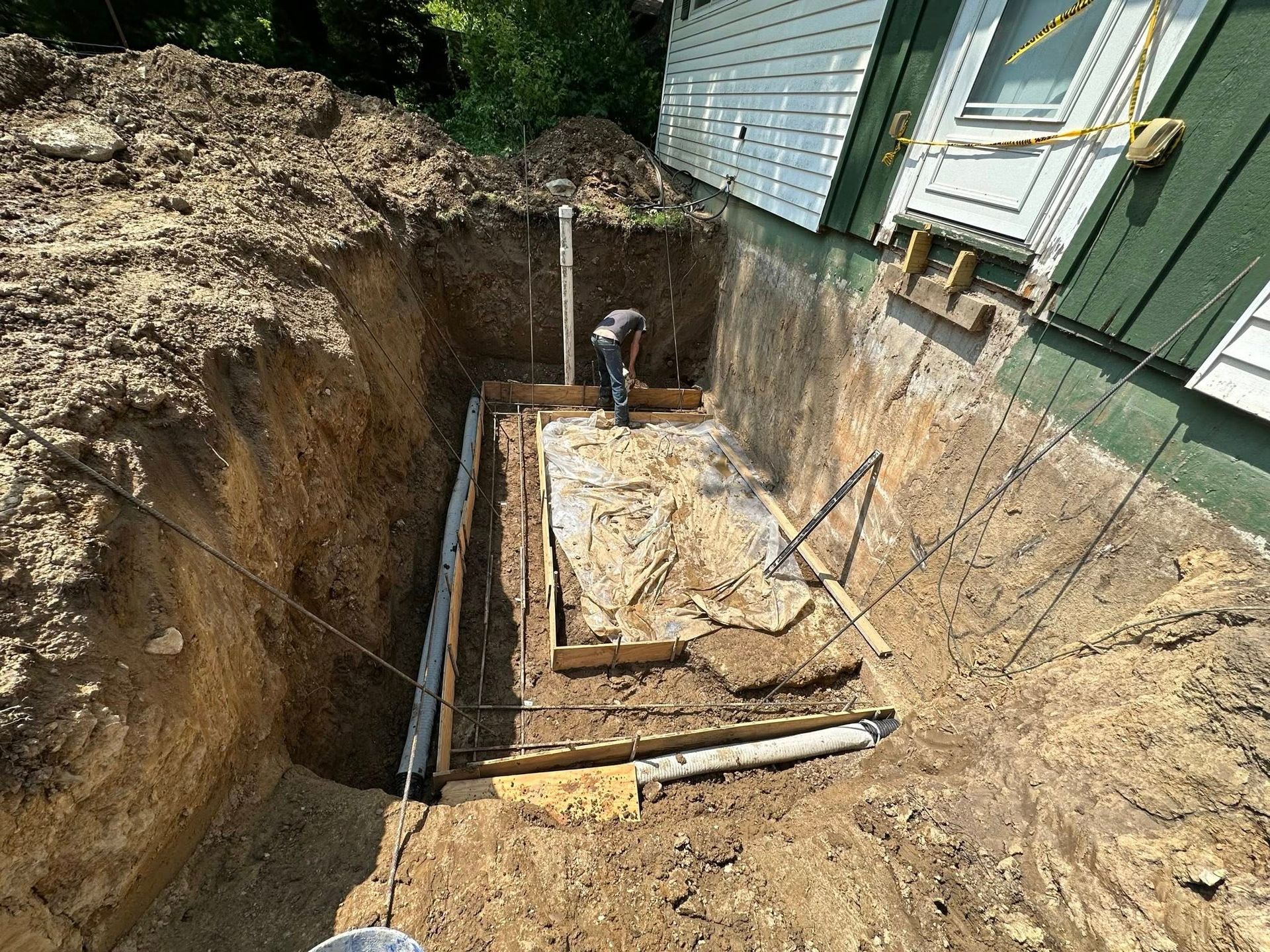 Excavation beside a house foundation with exposed soil, pipes, and a worker in a trench.