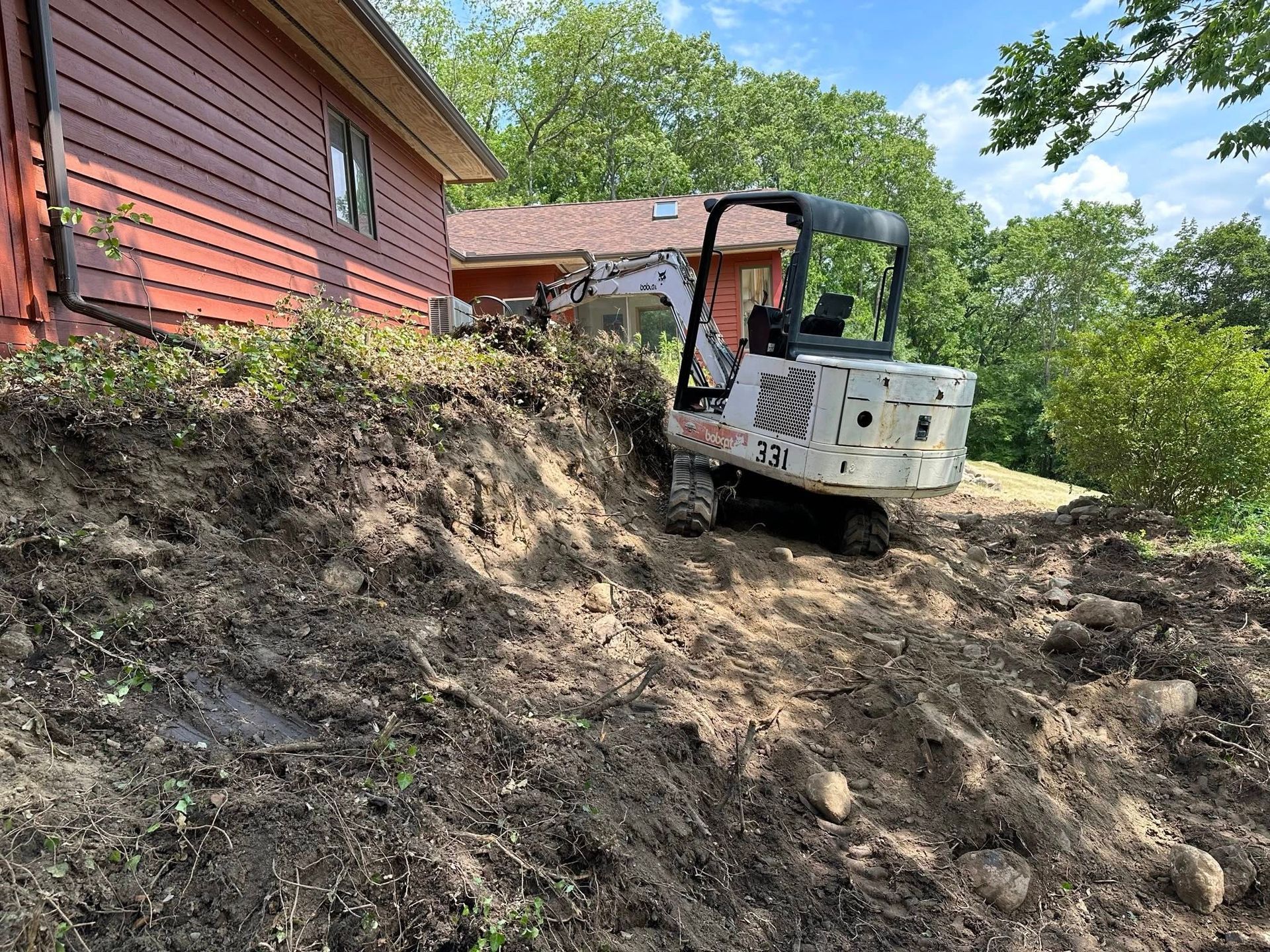 Small excavator on a dirt slope beside a red house, clearing soil near trees.