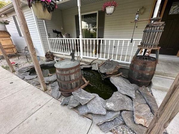 Front porch with two wooden barrels, stone steps, hanging flowers, and a small pond-like area in front