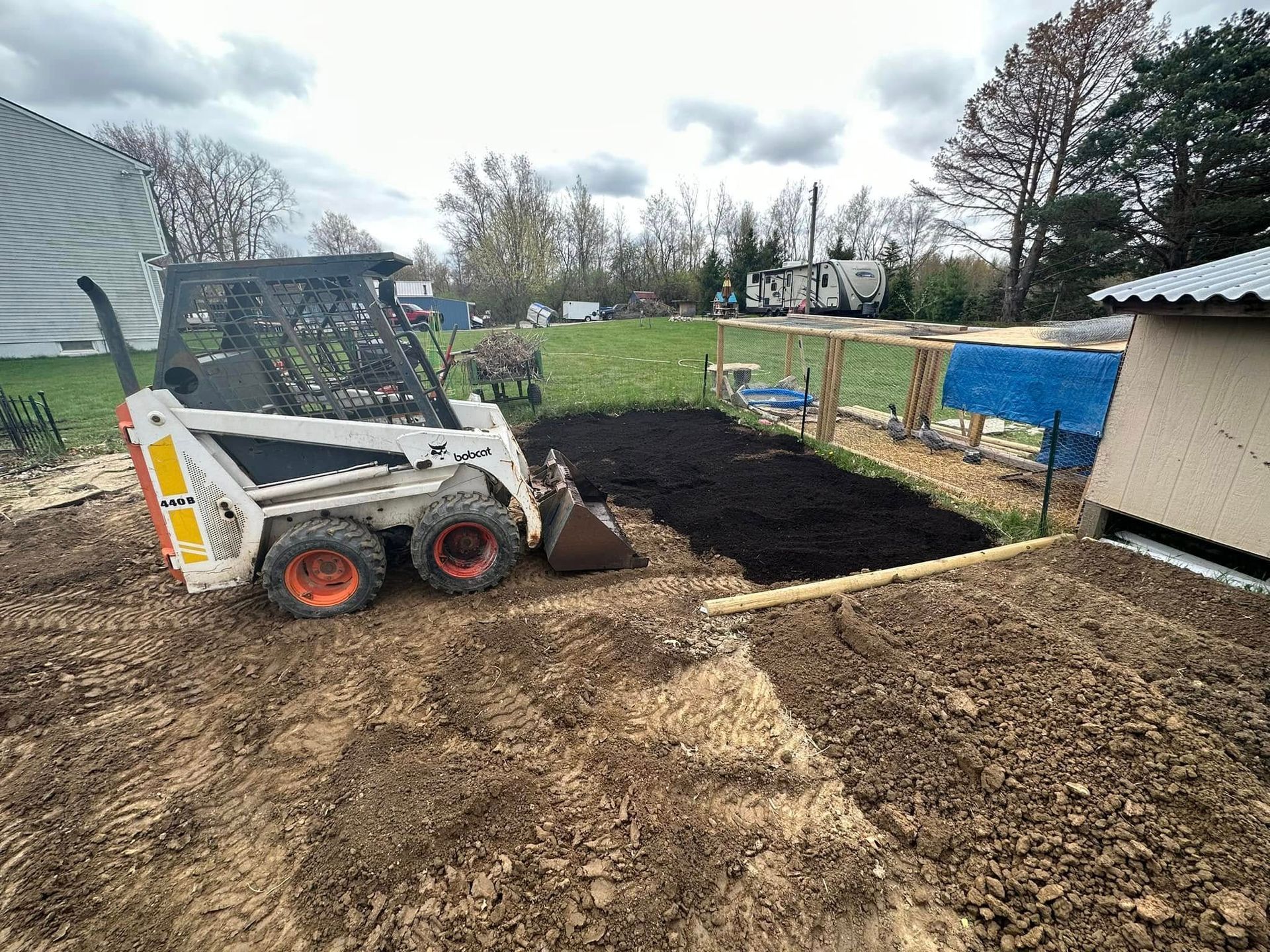 Skid steer spreading dark soil in a backyard beside a shed and fenced garden