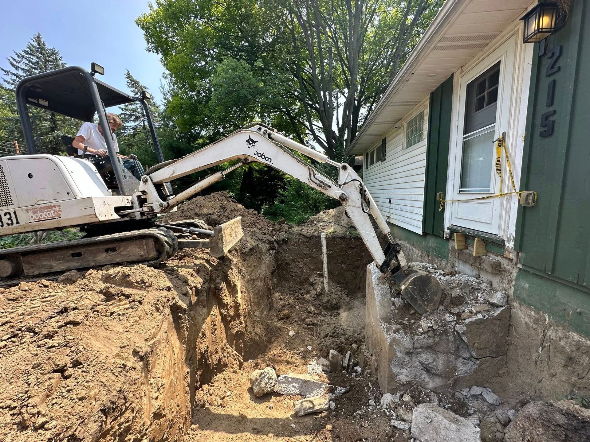 Excavator digging beside a house, removing soil near the foundation and front steps.