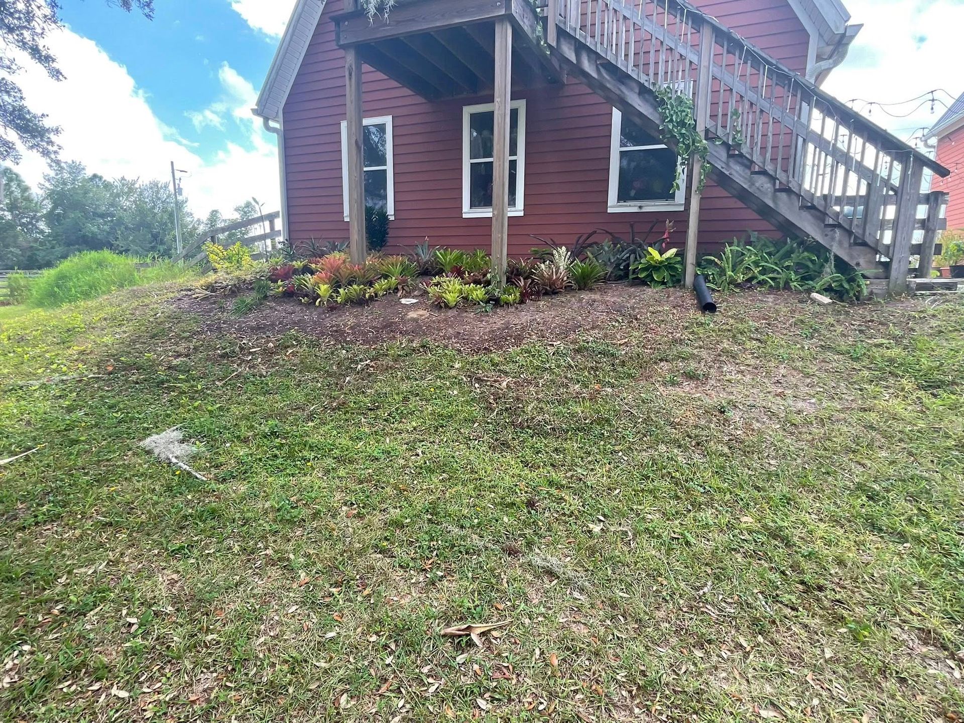 Red house with white trim on a grassy slope, front stairs and a flower bed under a cloudy sky