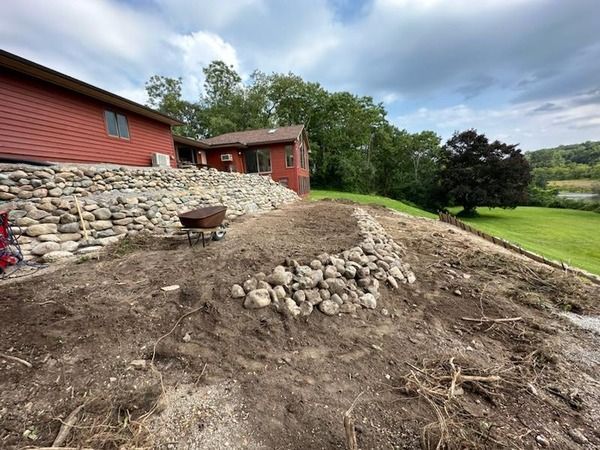 Red house beside a rocky, dirt-covered slope with trees and a green field beyond.