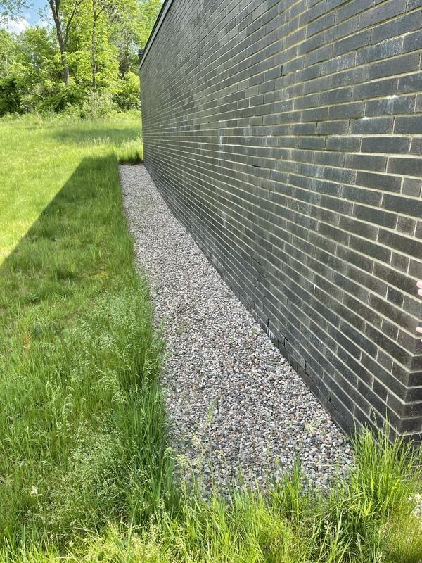 Gravel strip beside a dark brick wall, bordered by green grass and trees in sunlight.