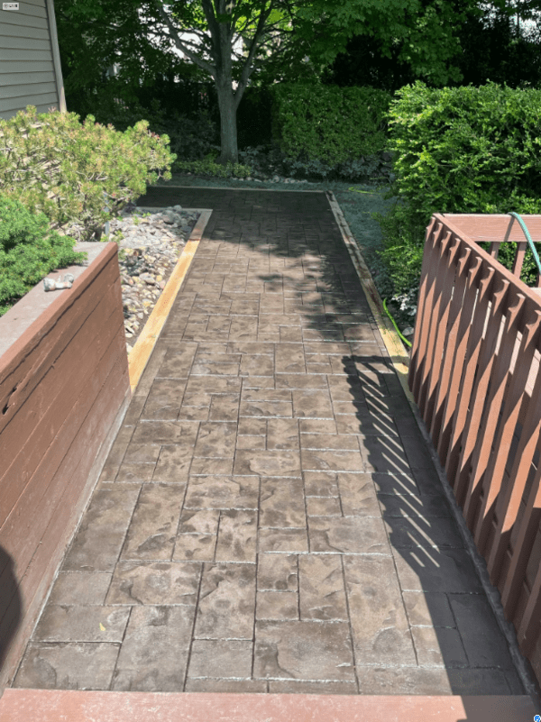 Brick walkway between wooden railings leading to a shaded garden path and greenery.