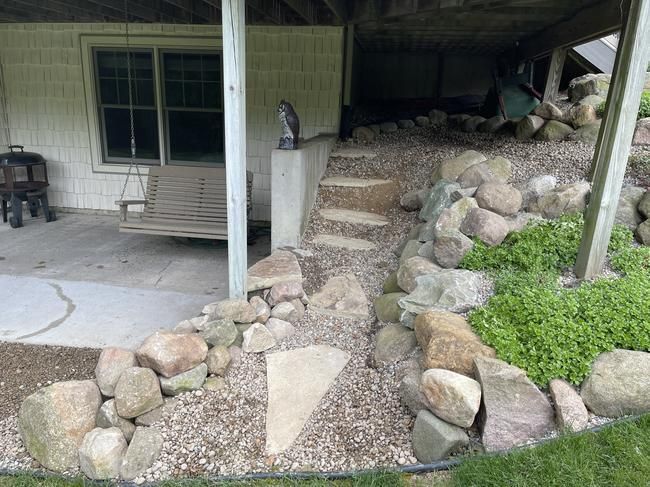 Stone steps and a gravel path leading up to a covered porch beside a house, bordered by large rocks and greenery