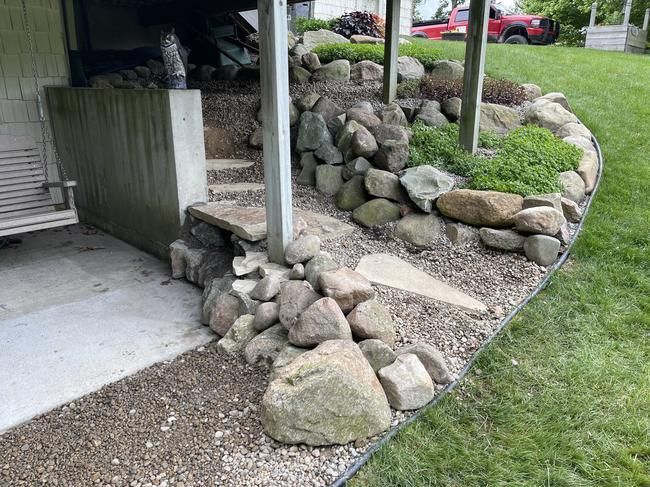 Rock-lined retaining wall and gravel bed beside a house porch, with lawn and parked cars in background