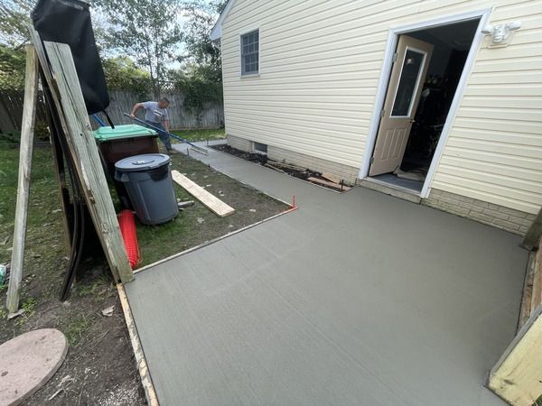 Freshly poured concrete patio beside a beige house with an open door and trash bins nearby