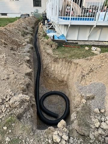 Black pipe laid in a trench beside a house foundation and porch under construction.