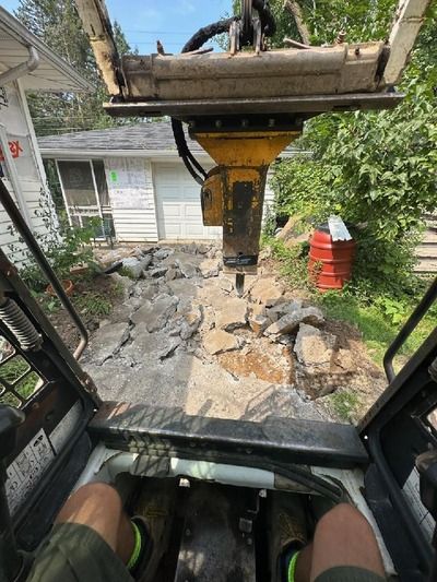 Inside an excavator cab, the bucket hovers over a torn-up backyard with debris and a red barrel.