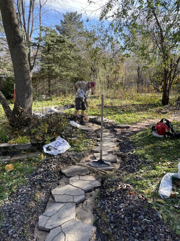 Person working in a wooded yard beside a stone path and garden tools