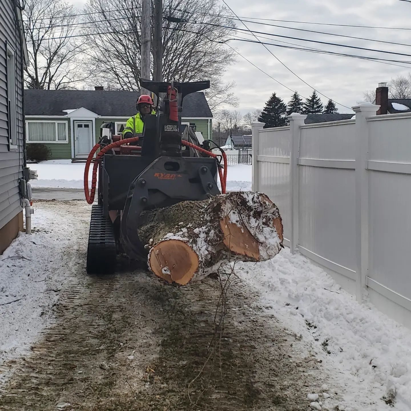A skid steer with a tree trunk in its grapple, moving down a snowy path beside a white fence.