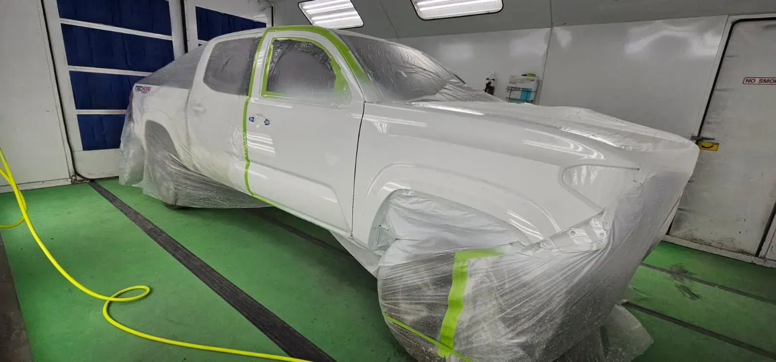 White pickup truck in a paint booth, covered in plastic and masking tape, ready for painting.