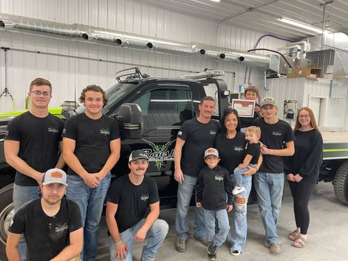 Group of people posing by a black truck in a garage; some are wearing matching shirts.