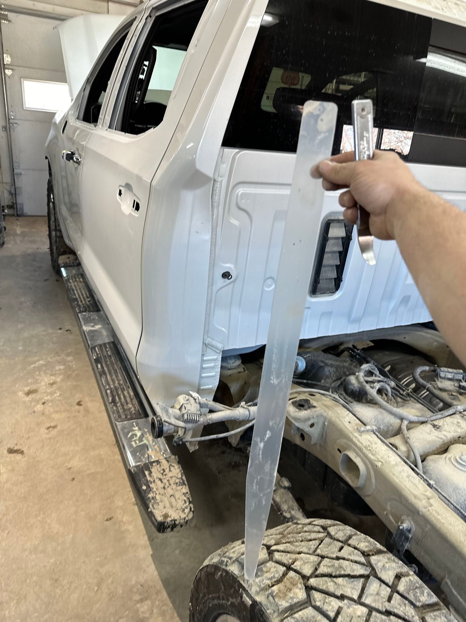 Person holds a metal measuring tool near a truck tire and vehicle body in a repair shop.