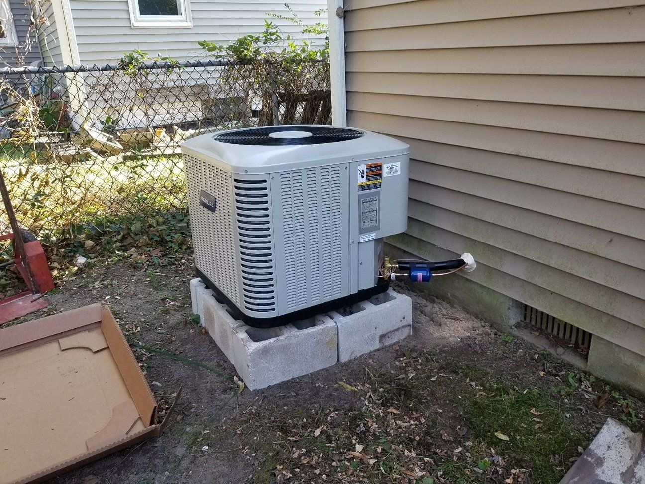 Outdoor air conditioner unit on concrete blocks beside a house wall, with cardboard and yard debris nearby