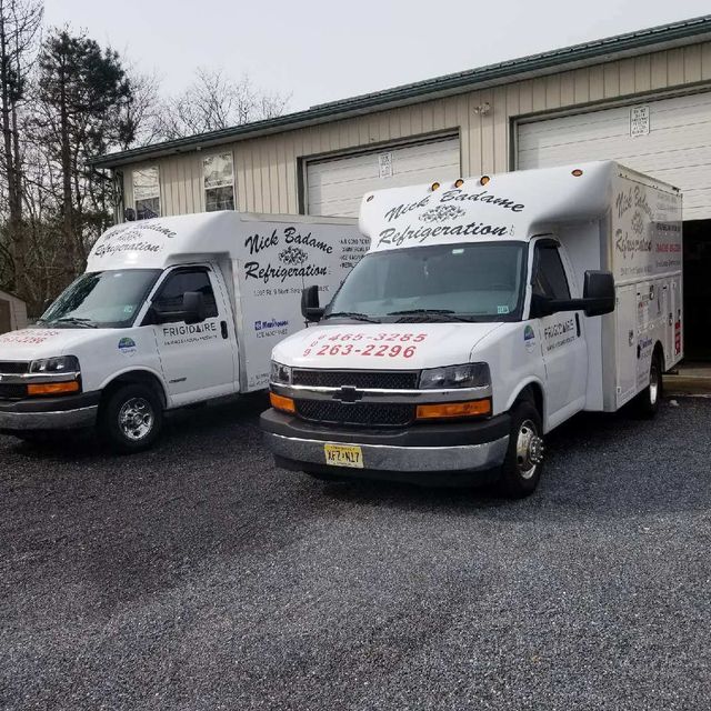 Two white utility vans with box bodies parked on gravel beside a warehouse.