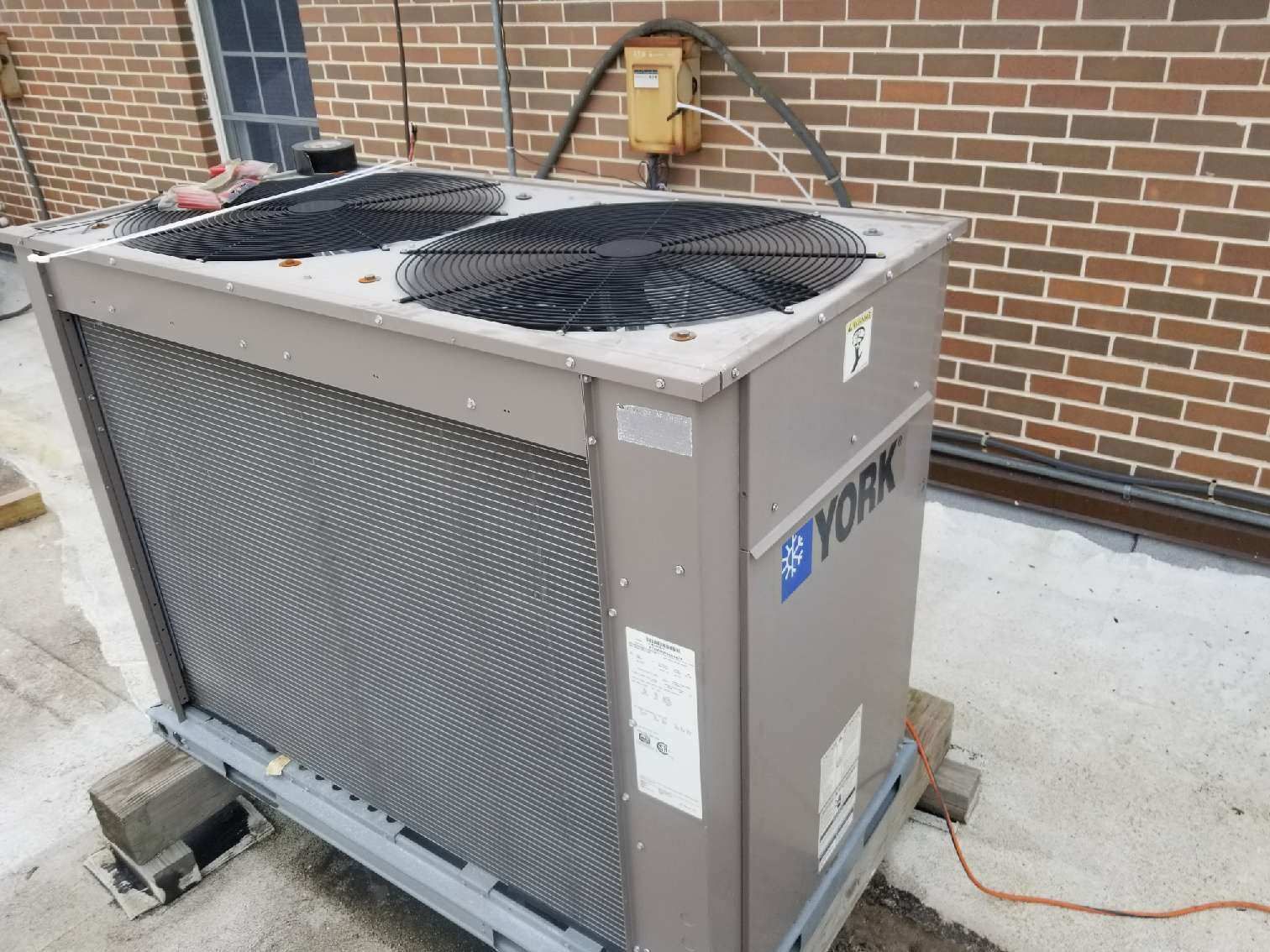Outdoor HVAC unit beside a brick wall, with a brick and concrete surface around it.