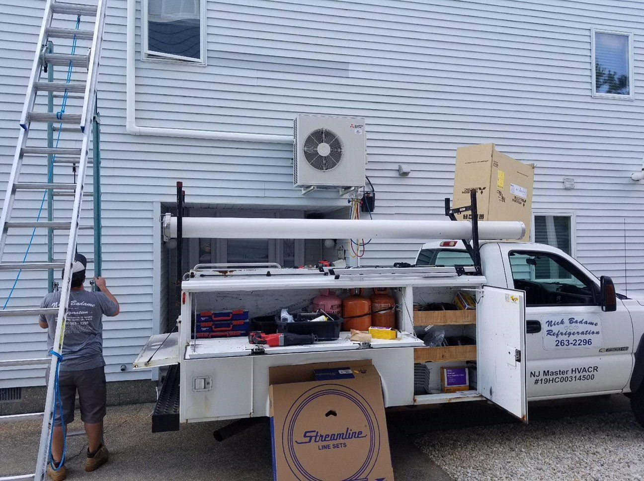 Utility truck with mounted equipment parked beside a house; worker stands near a ladder on the left.