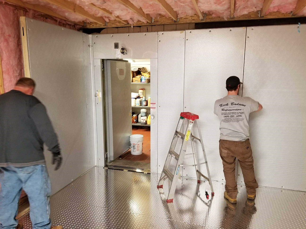 Two workers installing drywall in an unfinished room with exposed insulation and a ladder.