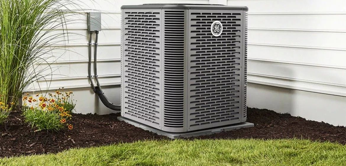 Gray outdoor air conditioner beside a house, next to a flower bed and green lawn