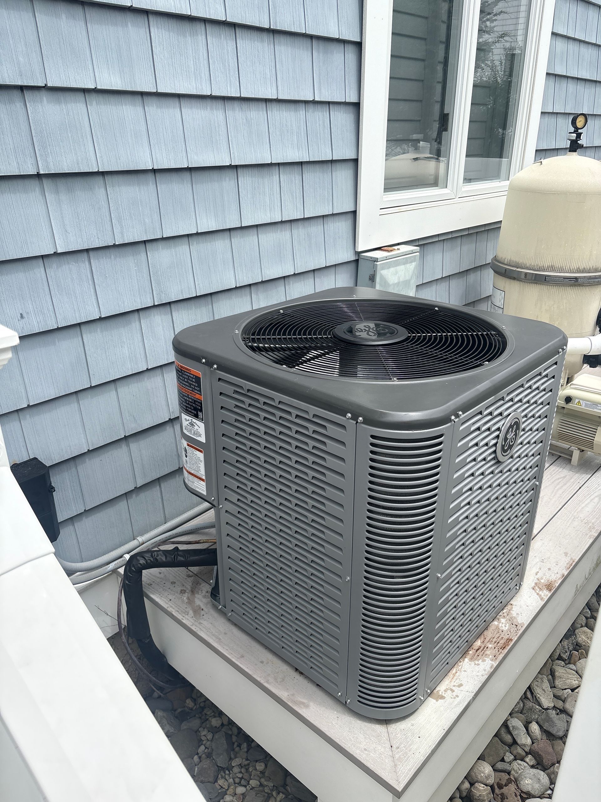 Outdoor air conditioner unit beside a gray house with windows and stone landscaping