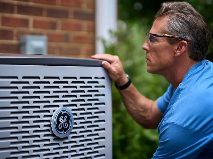 Technician inspecting a GE outdoor HVAC unit beside a brick wall