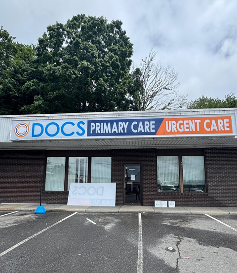 DOCS Primary Care Urgent Care storefront with sign, brick exterior, and empty parking lot under cloudy sky