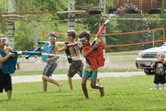 Children playing water guns on a grassy field; wearing vests and goggles, water spraying, sunny day.