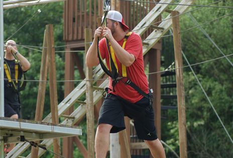 Man on zip line platform, wearing red shirt, holding ropes, wearing a safety harness.