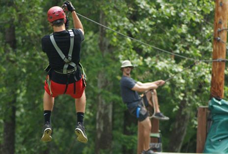 Man zip-lining, wearing helmet and safety harness. Another man waits. Forest background.