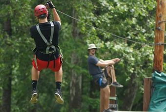 Man zip-lining, wearing helmet and safety harness. Another man waits. Forest background.