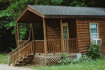 Cabin with wooden porch, door, and window. Log siding, brown roof, and greenery.