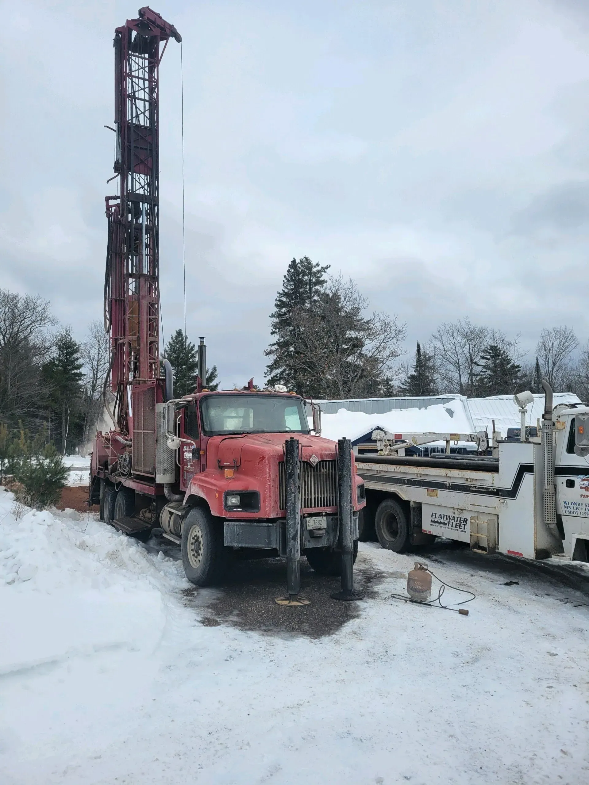 Red drilling rig truck in snow; another truck parked nearby. Cloudy sky.