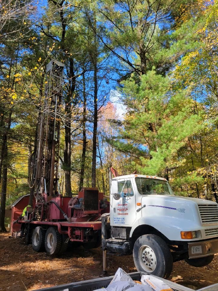 Drilling rig on a truck in a wooded area. White truck with red rig. Autumn leaves and blue sky.