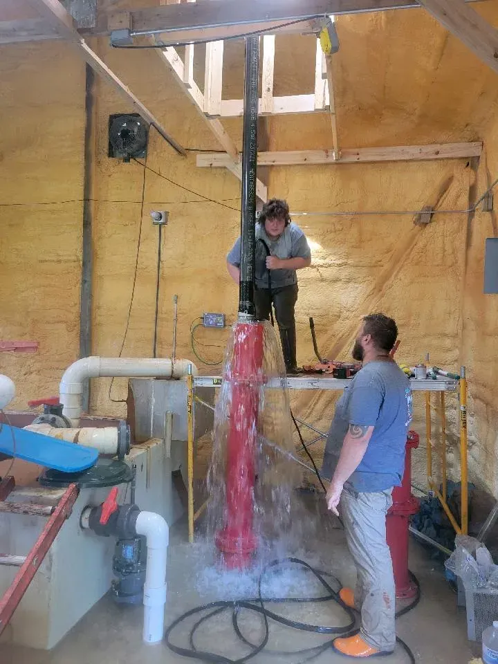 Two people work on a water pump, water sprays everywhere, inside a wooden structure.