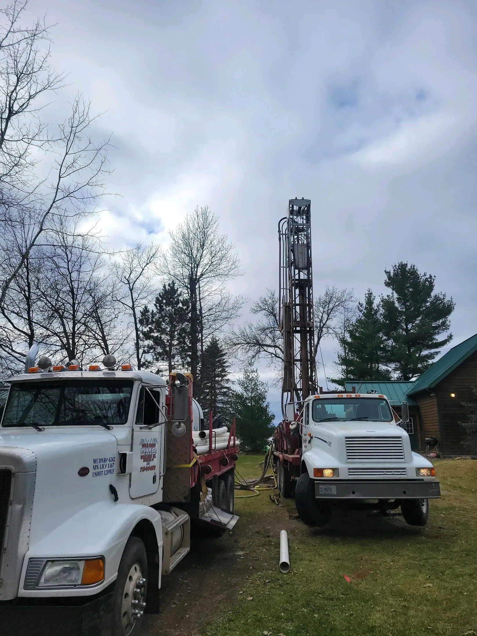 Two white well-drilling trucks parked near a cabin, cloudy sky above, surrounded by trees and grass.