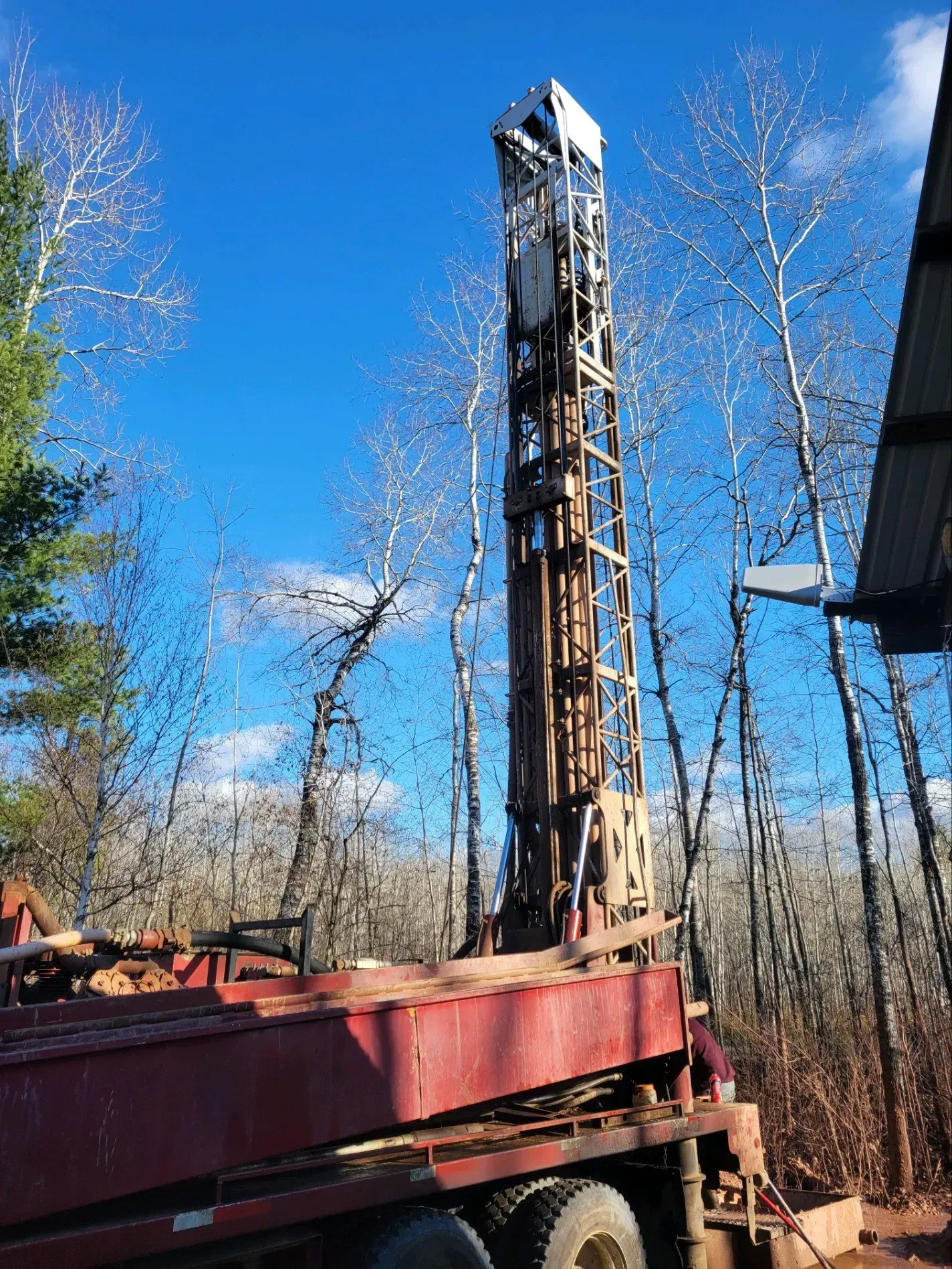 A red drilling rig on a truck under a blue sky with bare trees.