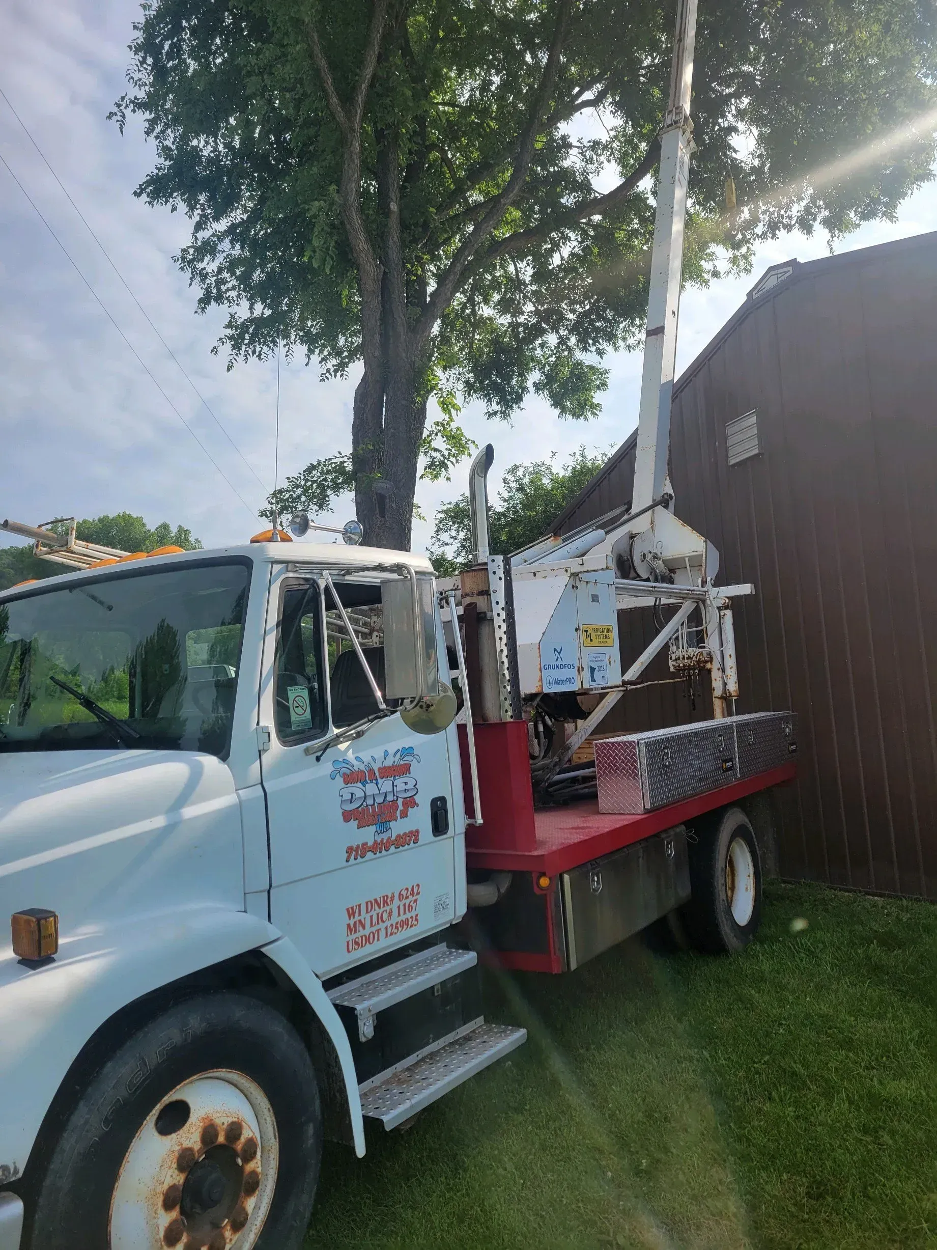 White utility truck with a raised lift arm, parked near a tree and brown shed.