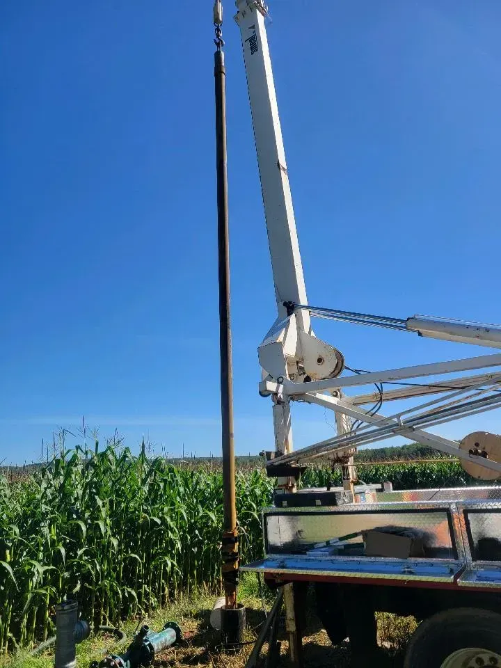 A well drilling rig with a tall, dark pipe, against a bright blue sky and green field.