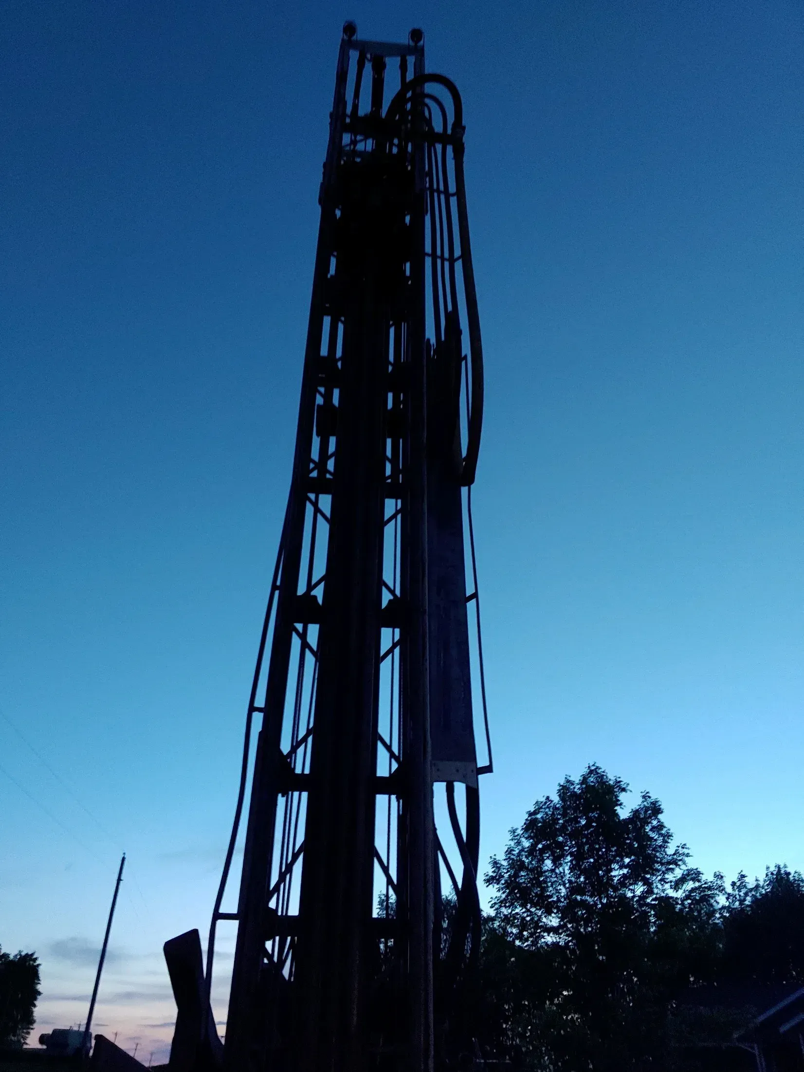 Silhouette of a drilling rig against a twilight blue sky, near trees.