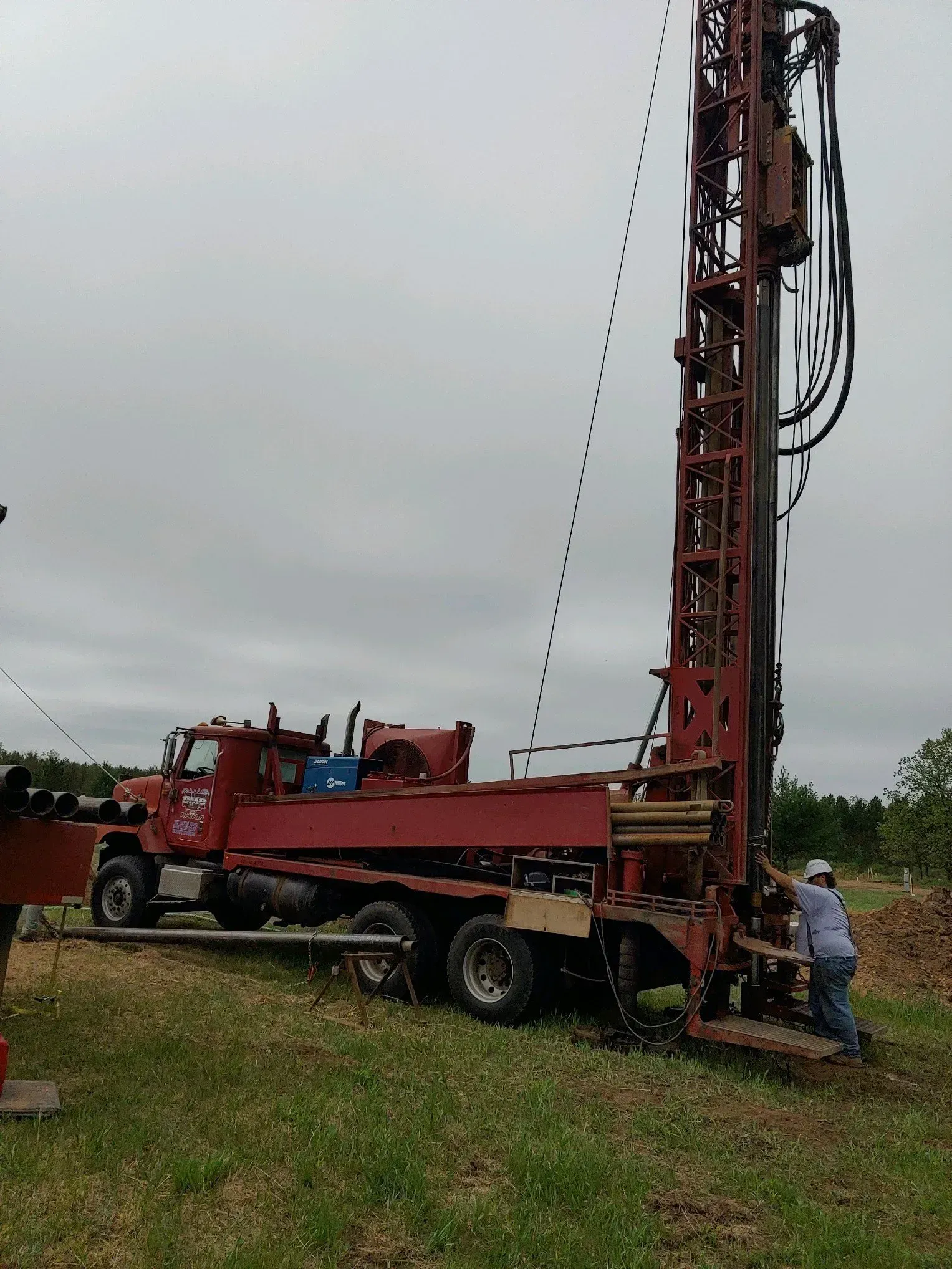 A red drilling rig on a truck, in a grassy field under a cloudy sky. A person is near the rig.