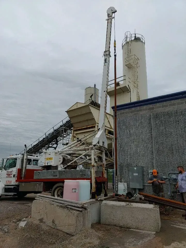 Truck-mounted crane next to industrial cement plant, with workers, cloudy sky.