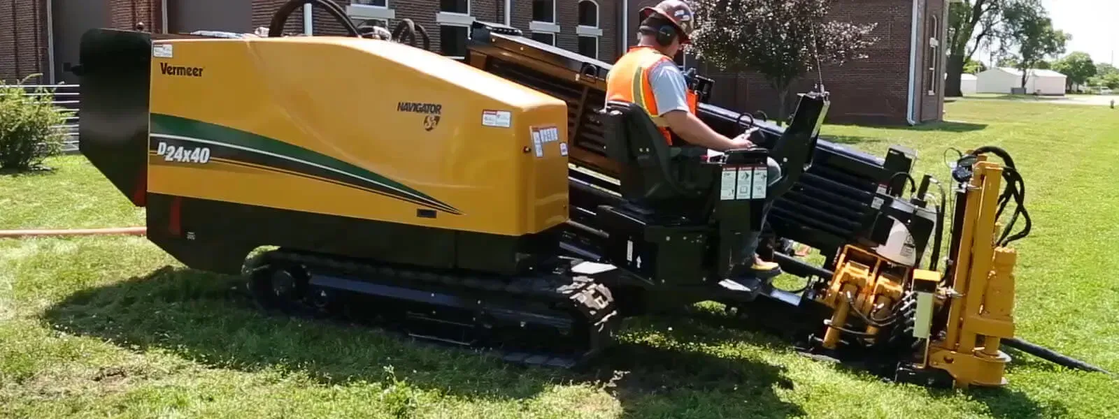 A worker operating a yellow drilling machine on grass. He wears a hard hat and safety vest.