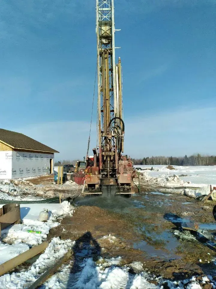 Well drilling rig at a construction site, with mud and snow, under a clear blue sky.