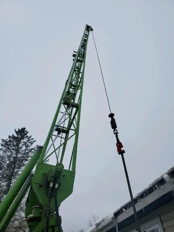 Green crane reaching towards a cloudy sky, hoisting a long, dark pole near a building.