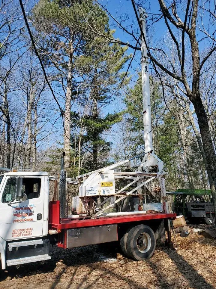 Truck-mounted drilling rig drilling in a wooded area. White tower, red flatbed, blue sky, and bare trees.