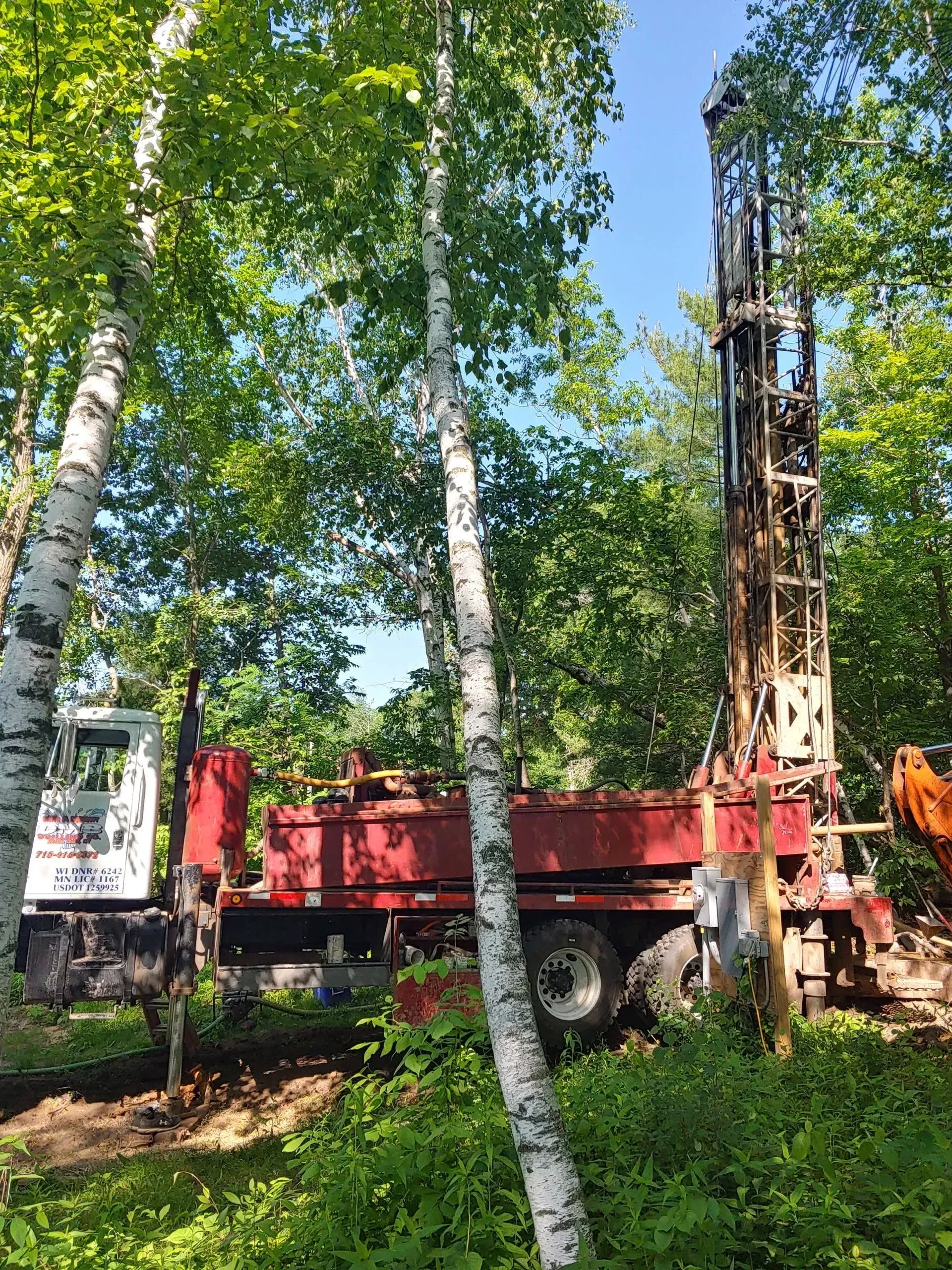 Drilling rig set up in a wooded area, red and white truck, trees surrounding.